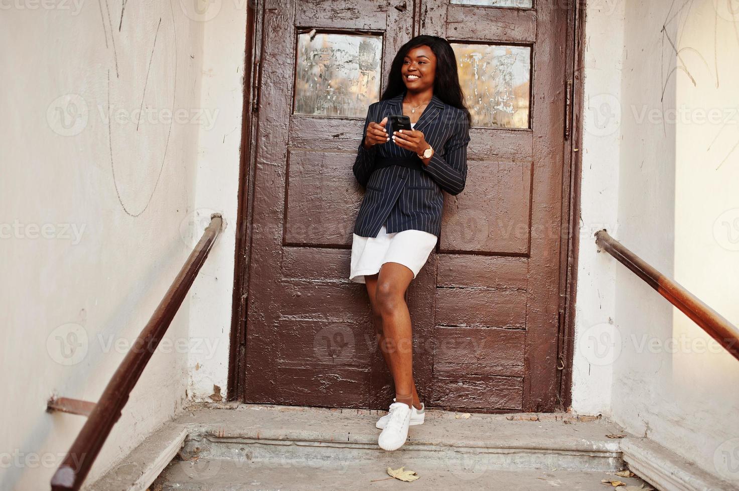 éxito elegante mujer afroamericana en chaqueta y falda contra la vieja puerta de madera con el teléfono inteligente en las manos. foto