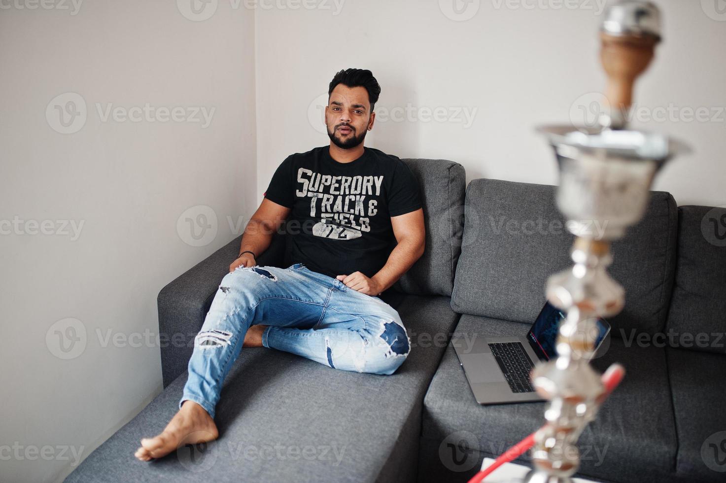 hombre indio de barba casual sentado en la habitación contra la cachimba con la computadora portátil. foto