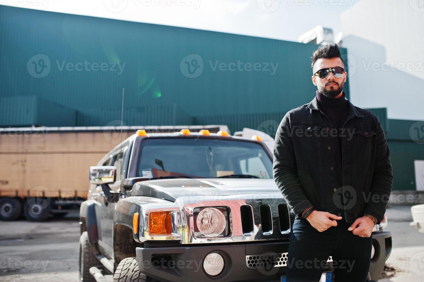 Fashion rich beard Arab man wear on black jeans jacket and sunglasses posed against big black suv car. Stylish, succesful and fashionable arabian model guy. photo
