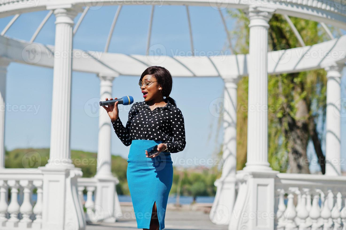 Stylish african american model girl TV presenter with microphone in glasses, blue skirt and black blouse posed outdoor against white stone arch with mobile phone. photo