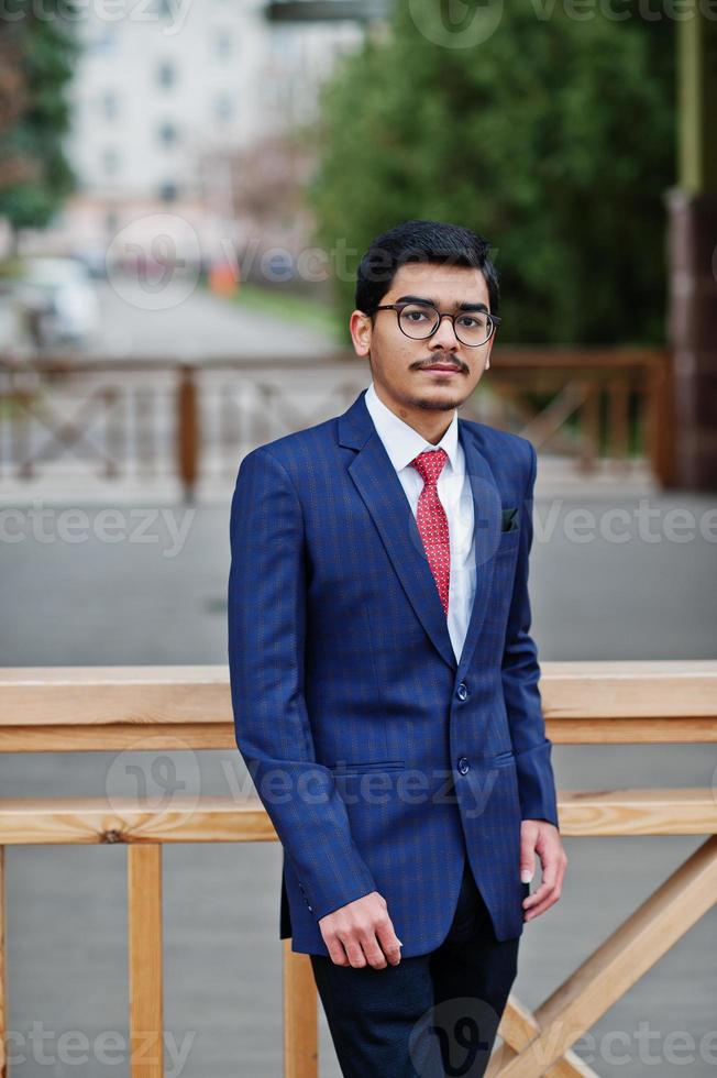 joven indio con anteojos, vestido con traje y corbata roja posado al aire libre. foto