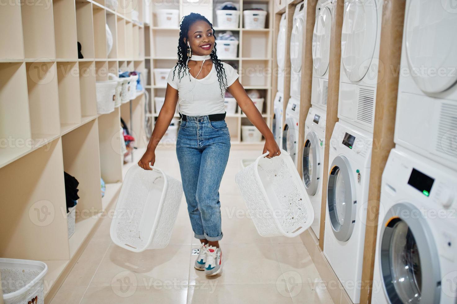 Cheerful african american woman near washing machine listening music by earphones from mobile phone in the self-service laundry. photo