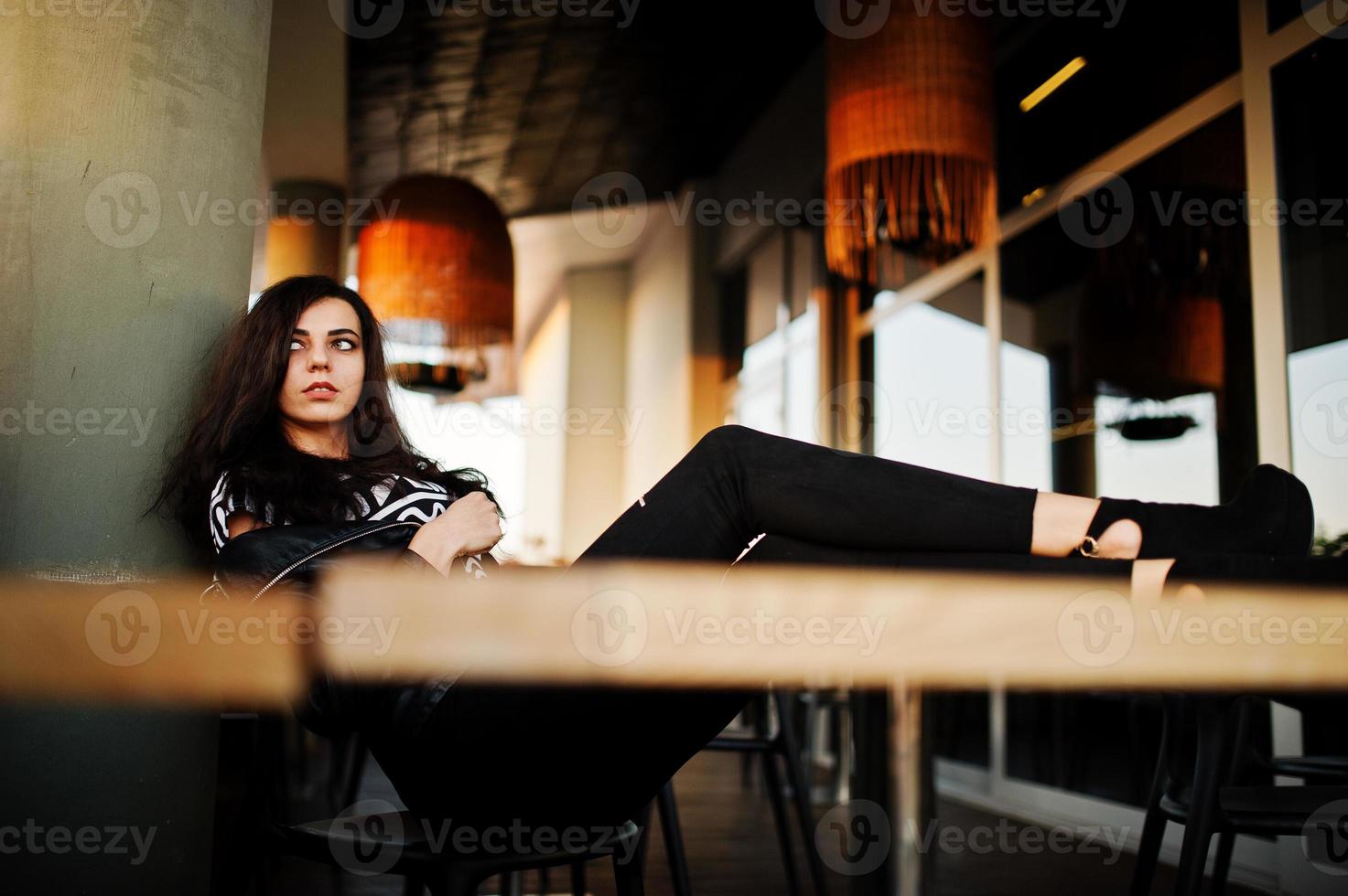 joven mujer rizada con chaqueta de cuero en un bar puso las piernas sobre la mesa. foto