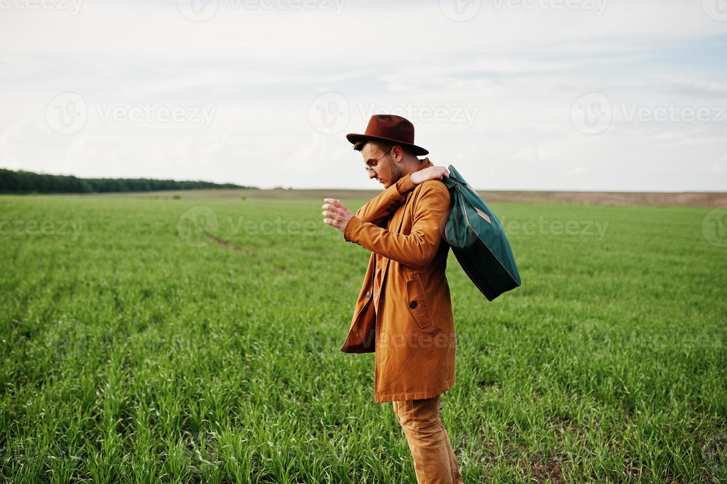 Stylish man in glasses, brown jacket and hat with bag posed on green field. photo
