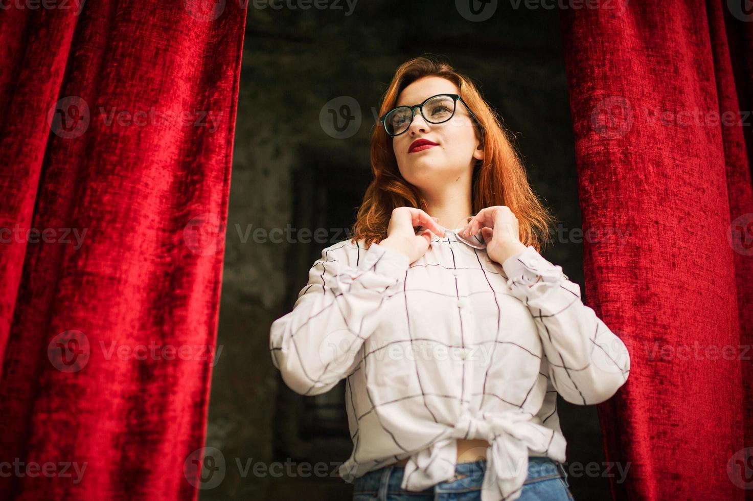 Attractive redhaired woman in eyeglasses, wear on white blouse posing at arch of open red curtains. photo