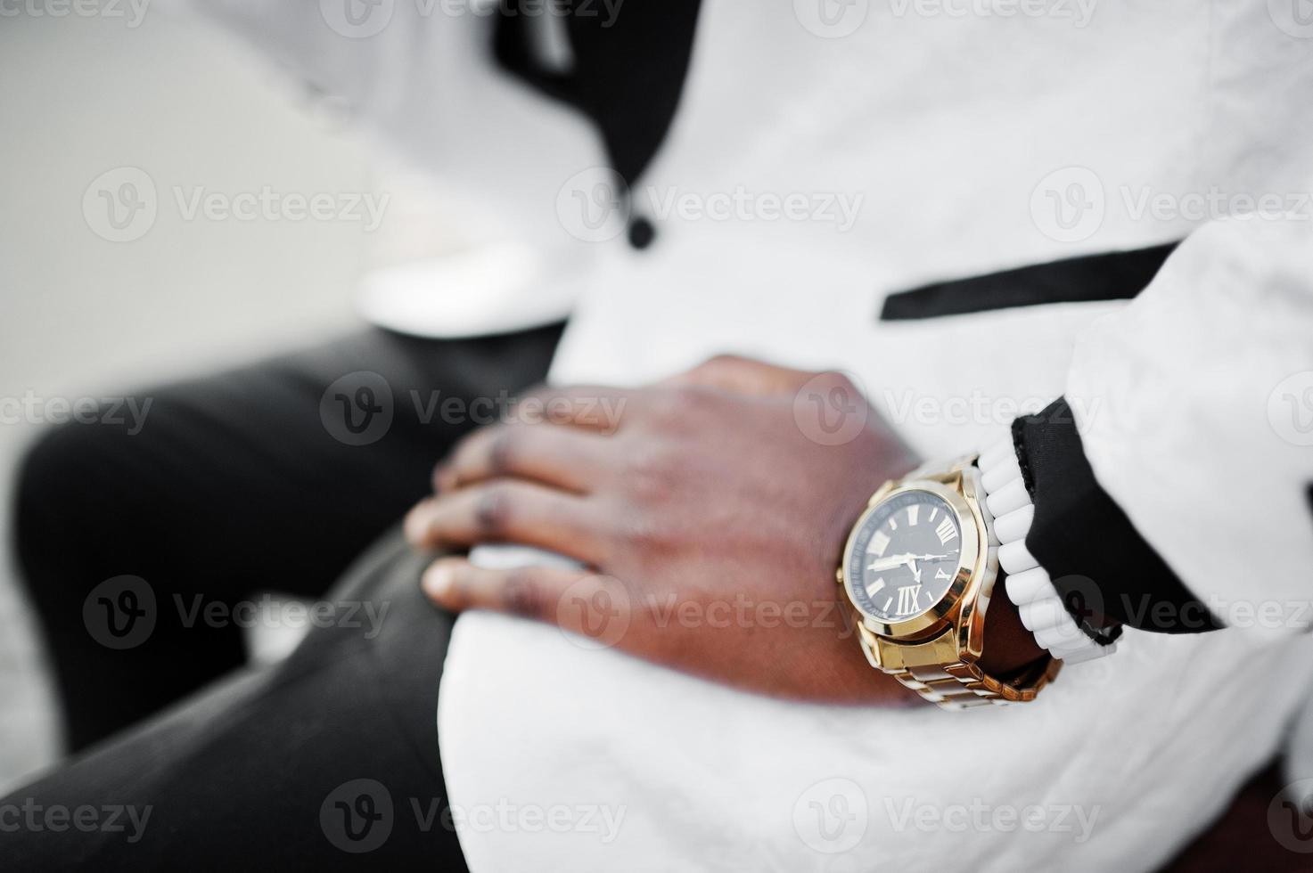 Chic handsome african american man in white suit sitting on bench. Close up photo of golden watches on hand.