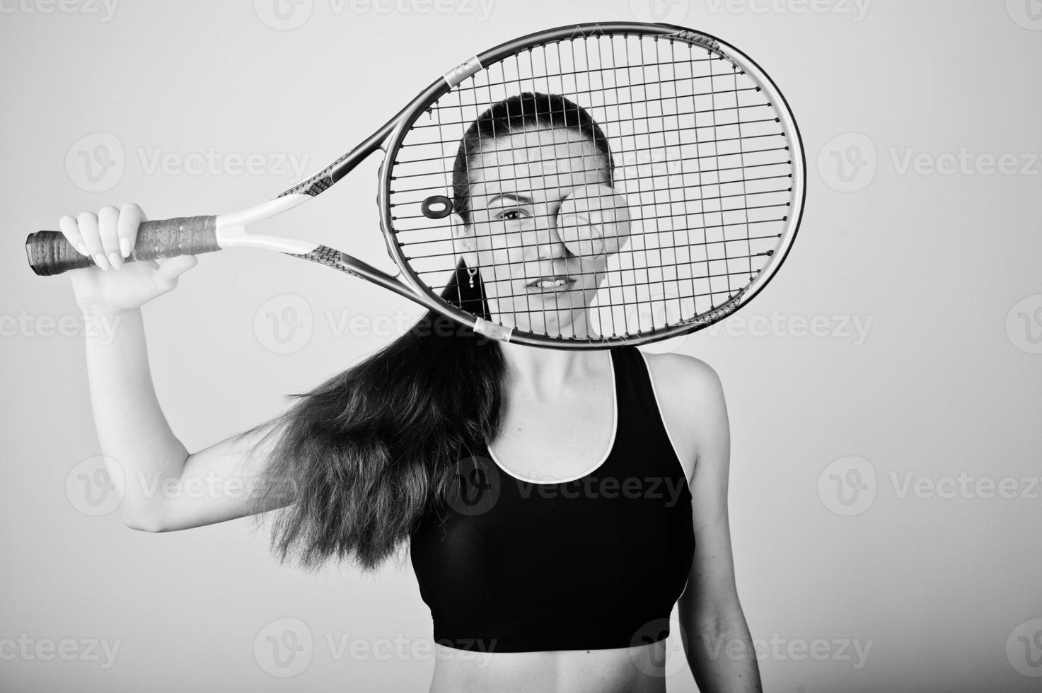 Black and white portrait of beautiful young woman player in sports clothes holding tennis racket while standing against white background. photo