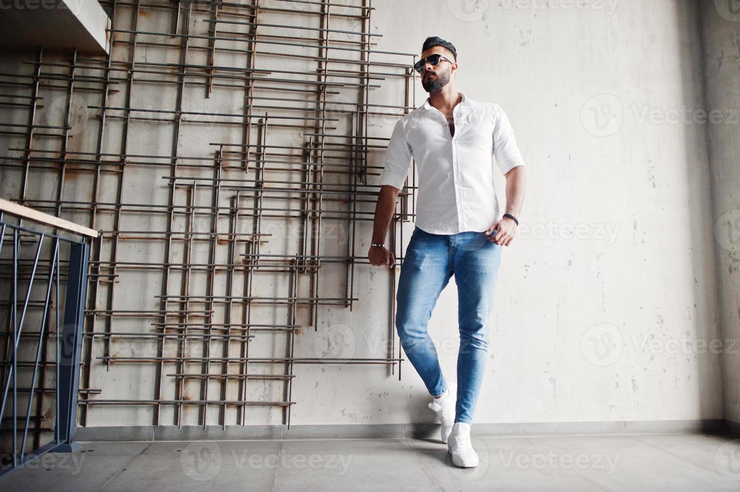 elegante modelo árabe alto con camisa blanca, jeans y gafas de sol posados contra una pared de acero en el interior. barba chico árabe atractivo. foto