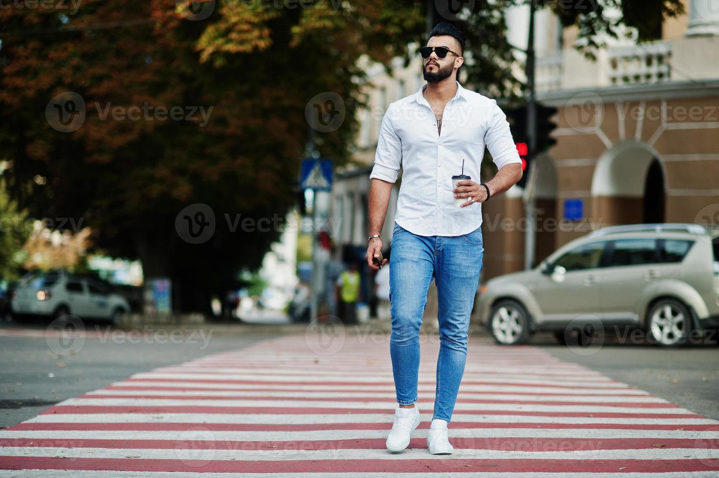 Stylish tall arabian man model in white shirt, jeans and sunglasses posed at street of city. Beard attractive arab guy with cup of coffee walking on pedestrian crossing or crosswalk. photo