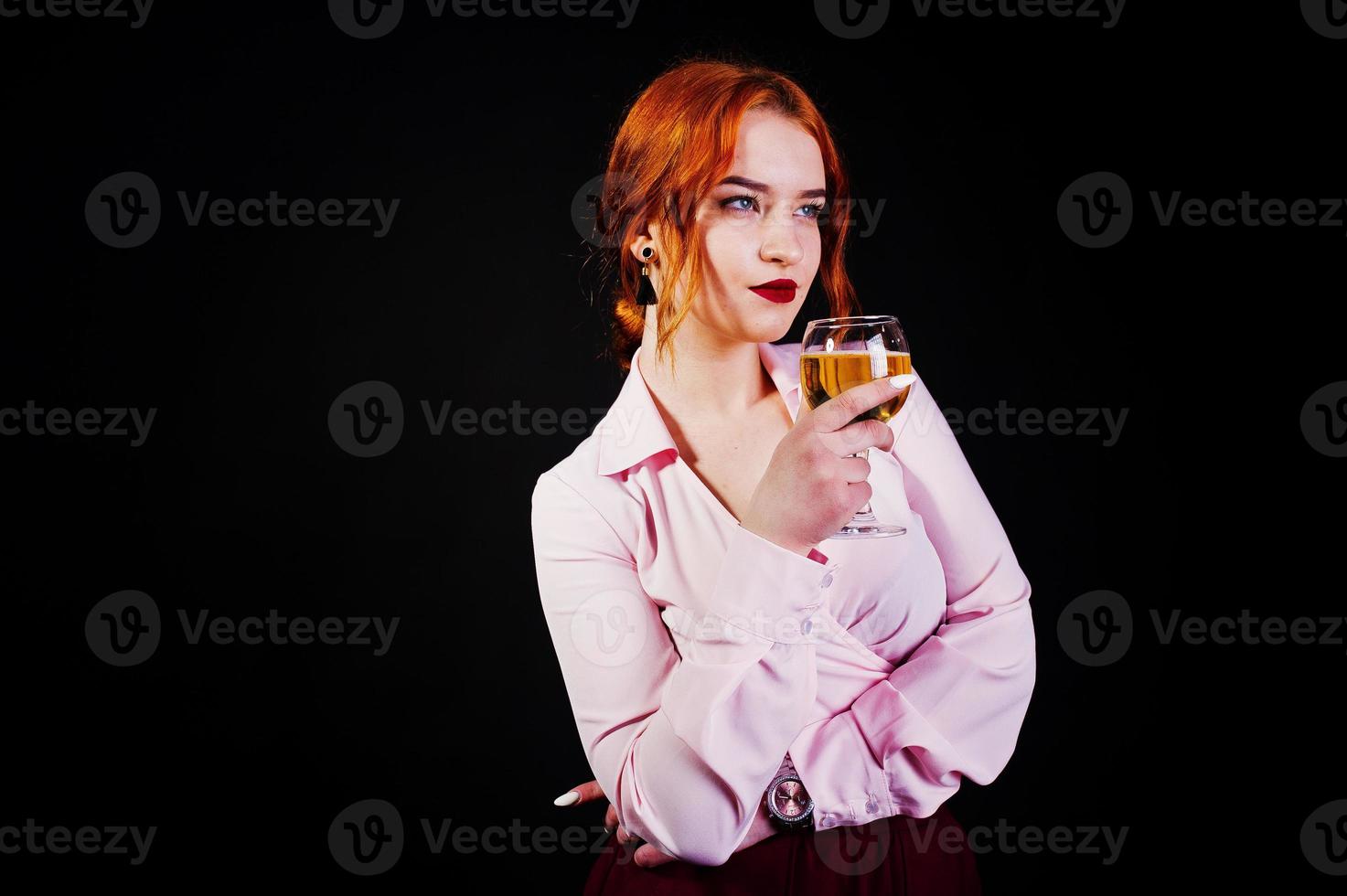 Gorgeous red haired girl in pink blouse and red skirt with glass of wine at hand isolated on black. photo