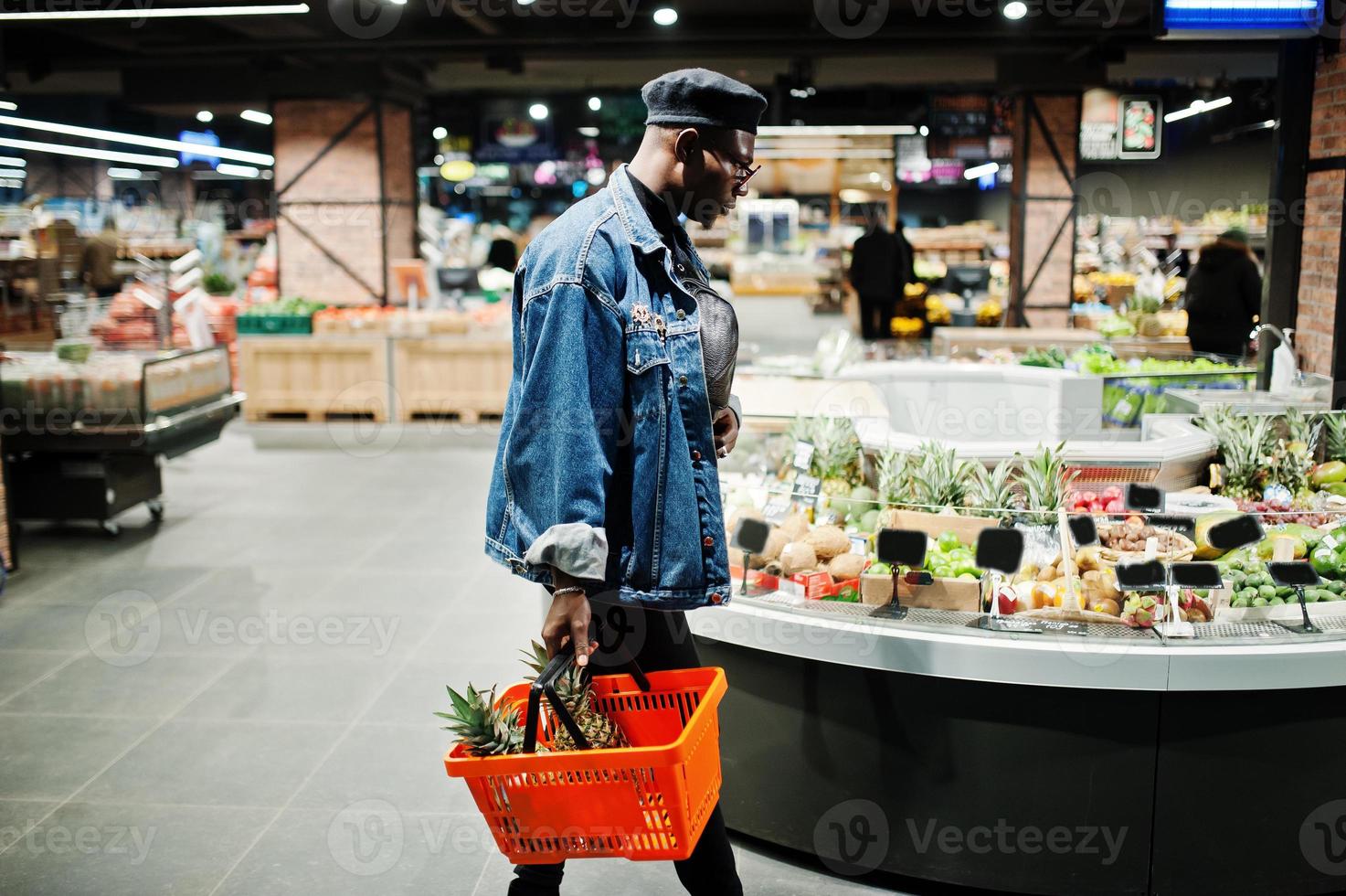 Elegante hombre afroamericano casual con chaqueta de jeans y boina negra sosteniendo una canasta con piñas en la sección orgánica de frutas del supermercado. foto