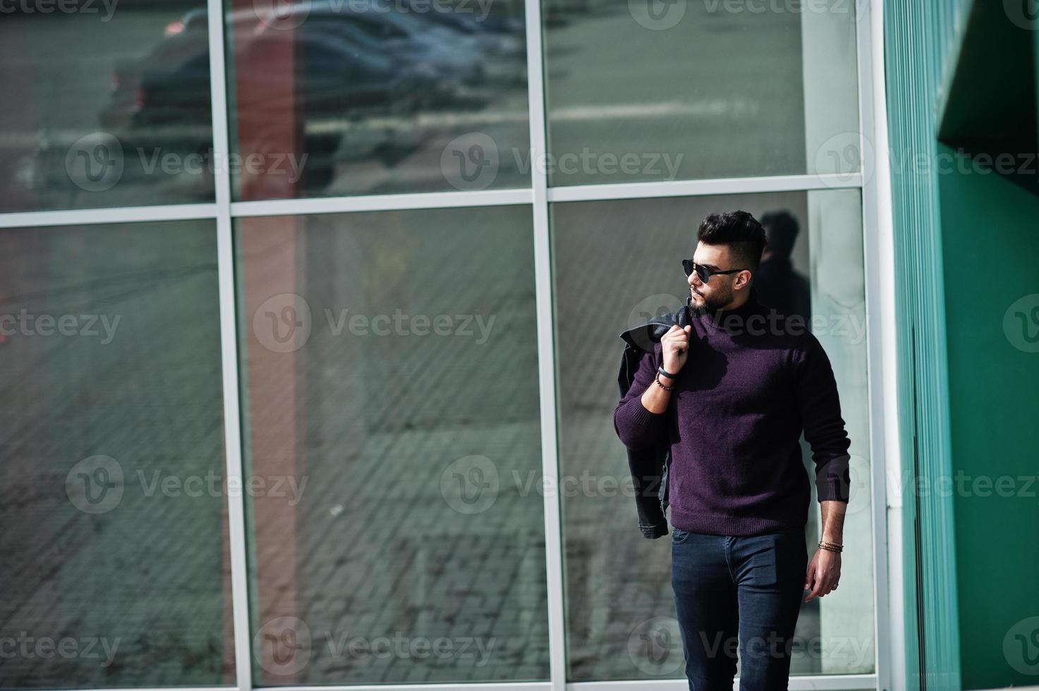 Fashion rich beard Arab man wear on turtle neck and sunglasses posed against large windows of modern building with jacket on hand. Stylish, succesful and fashionable arabian model guy. photo