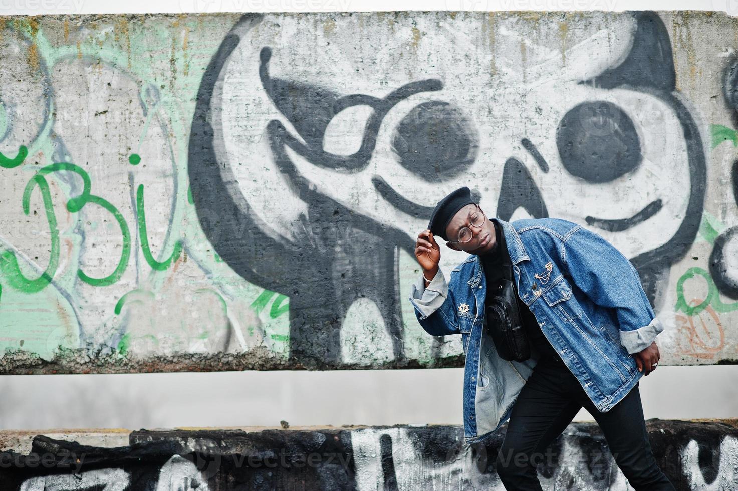 African american man in jeans jacket, beret and eyeglasses against graffiti wall with skull. photo