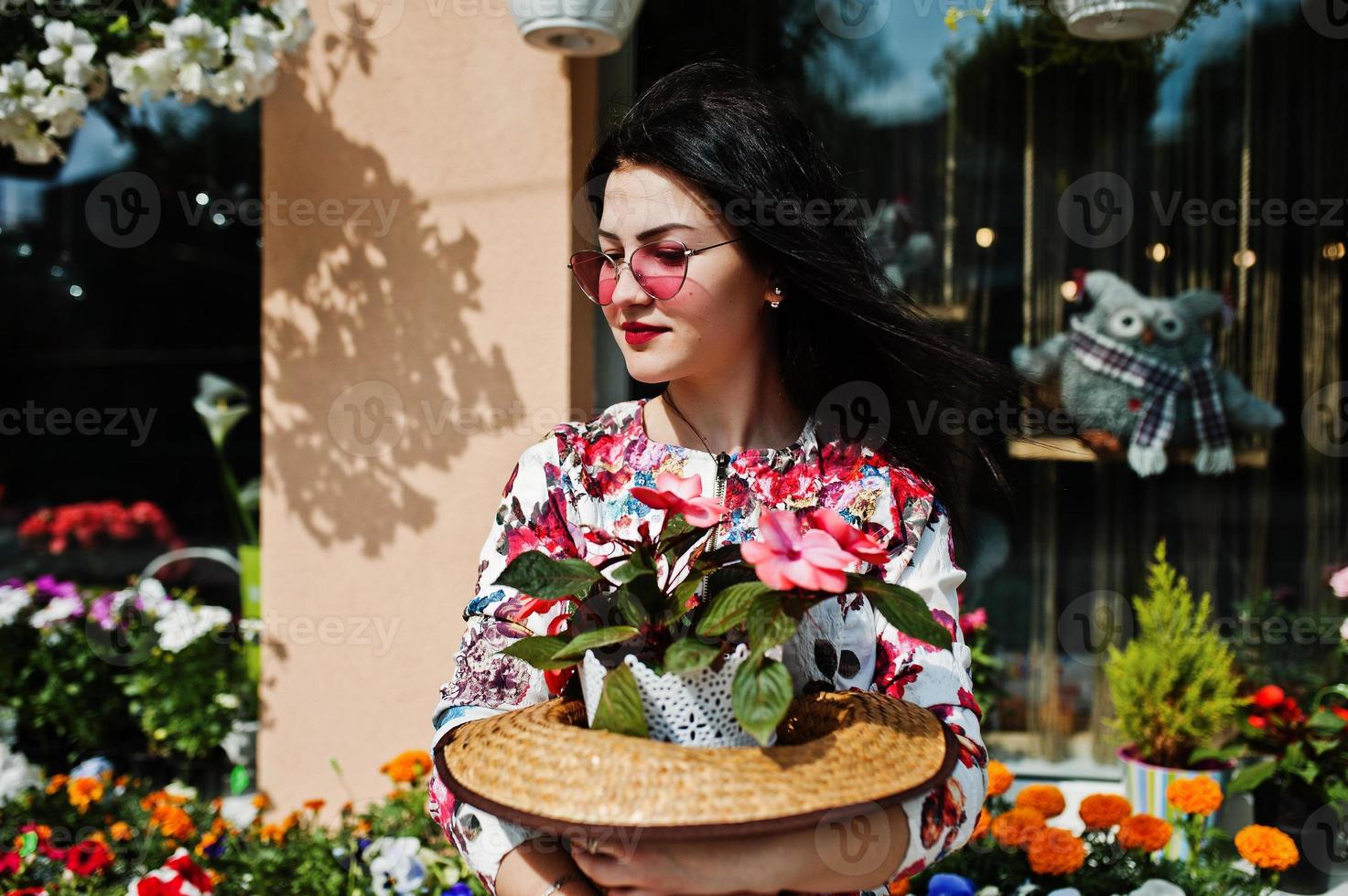 retrato de verano de una chica morena con gafas rosas y sombrero contra la tienda de flores. foto