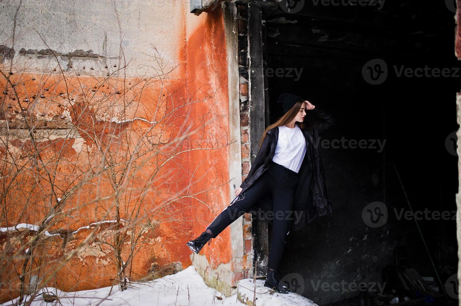 modelo morena de piernas largas de moda en un largo manto negro posado al aire libre en el día de invierno contra la vieja pared del grunge. foto