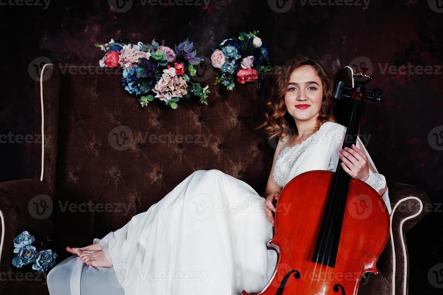 Pretty young gilrl musician in white dress with double bass sitting on brown vintage sofa. photo