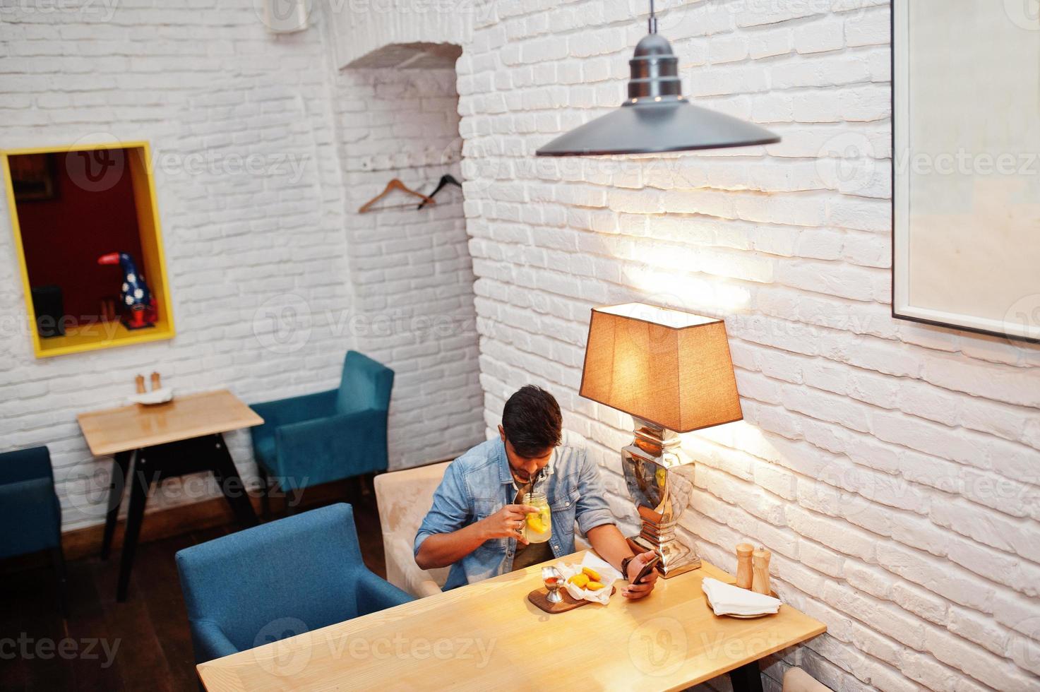 Portrait of handsome successful bearded south asian, young indian freelancer in blue jeans shirt sitting in cafe with chicken nuggets and lemonade. Hold mobile phone at hand. photo