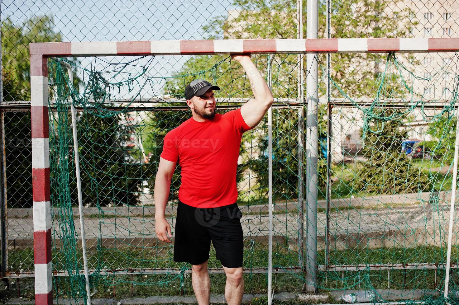 Young brutal bearded muscular man wear on red shirt, shorts and cap at stadium. photo