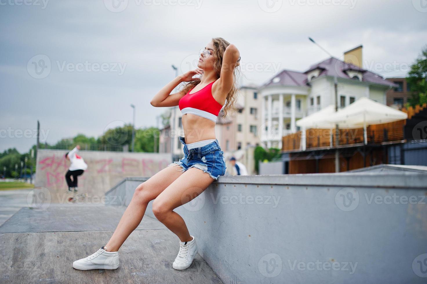Sexy curly model girl in red top, jeans denim shorts, eyeglasses and sneakers posed at skatepark. photo