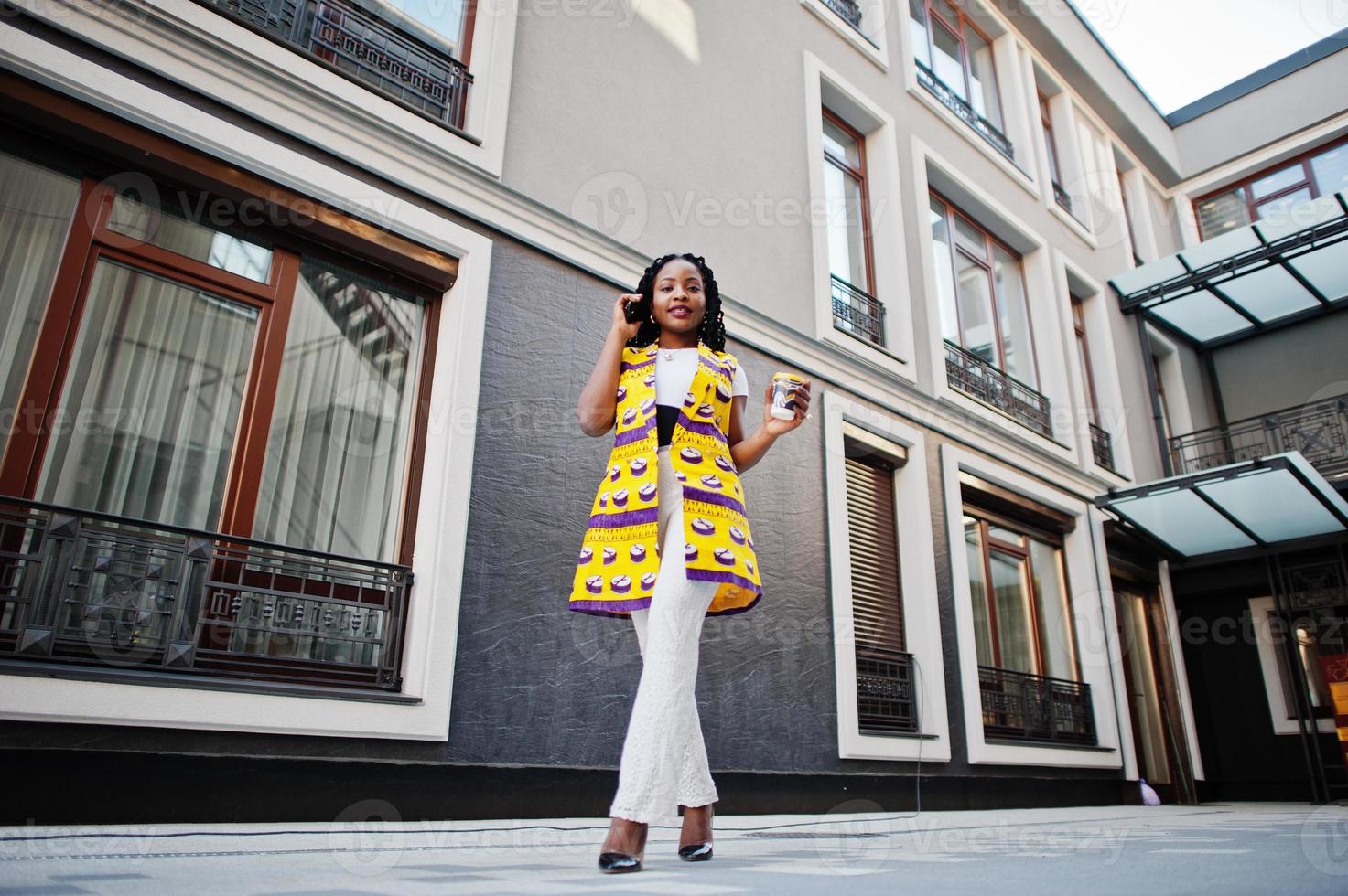 Stylish african american women in yellow jacket posed on street with hot drink in disposable paper cup and mobile phone at hands. photo