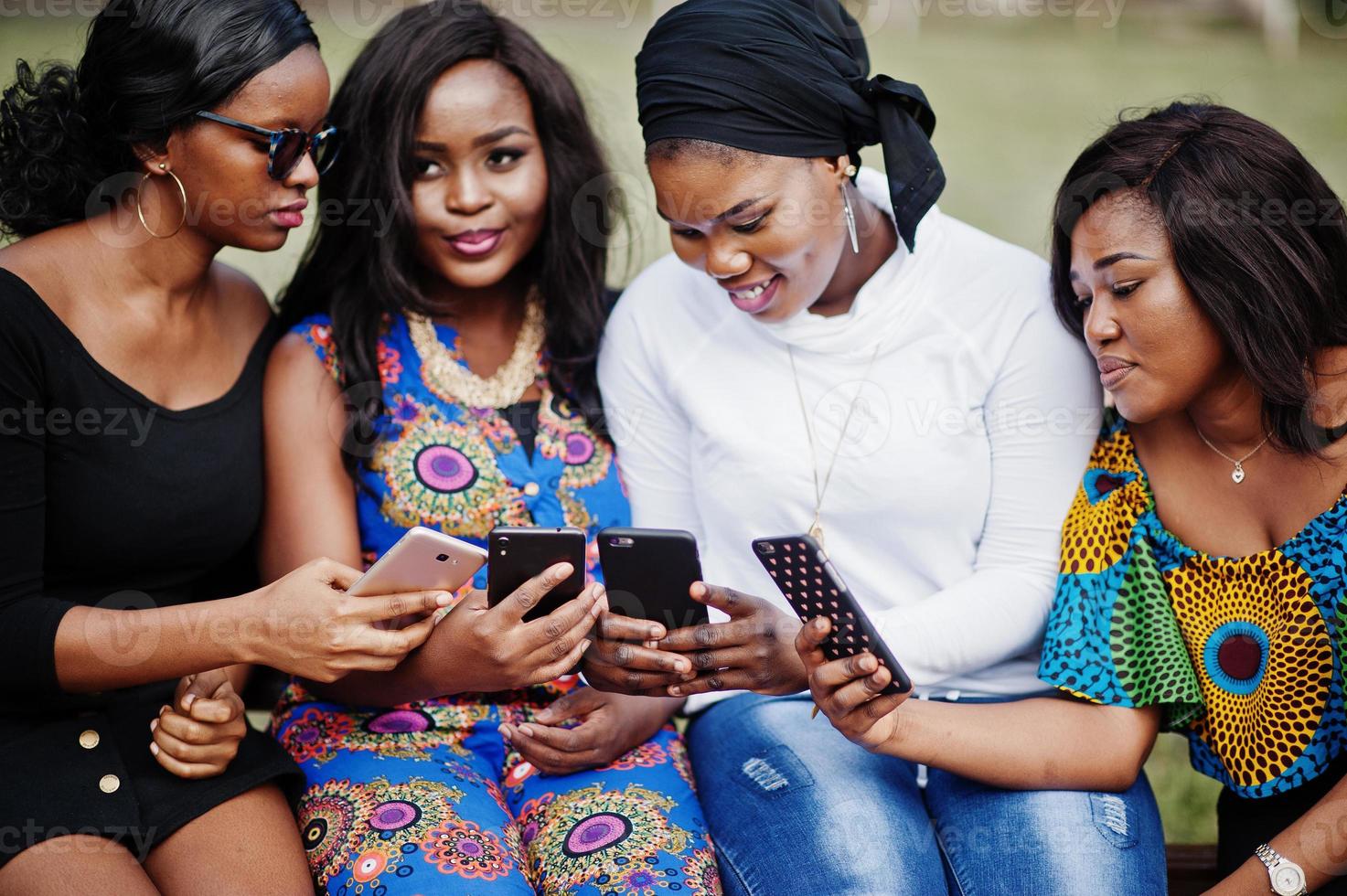 Group of four african american girls sitting on bench outdoor and looking at mobile phones. photo
