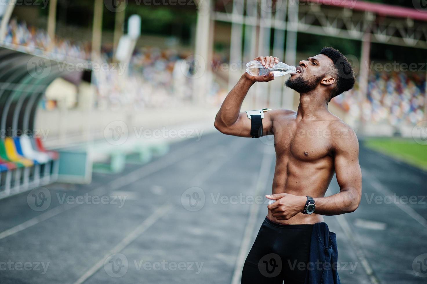 African american male athlete sexy sport bare torso man with running sports arm case for mobile phone, posed at stadium and drinking water from bottle. photo