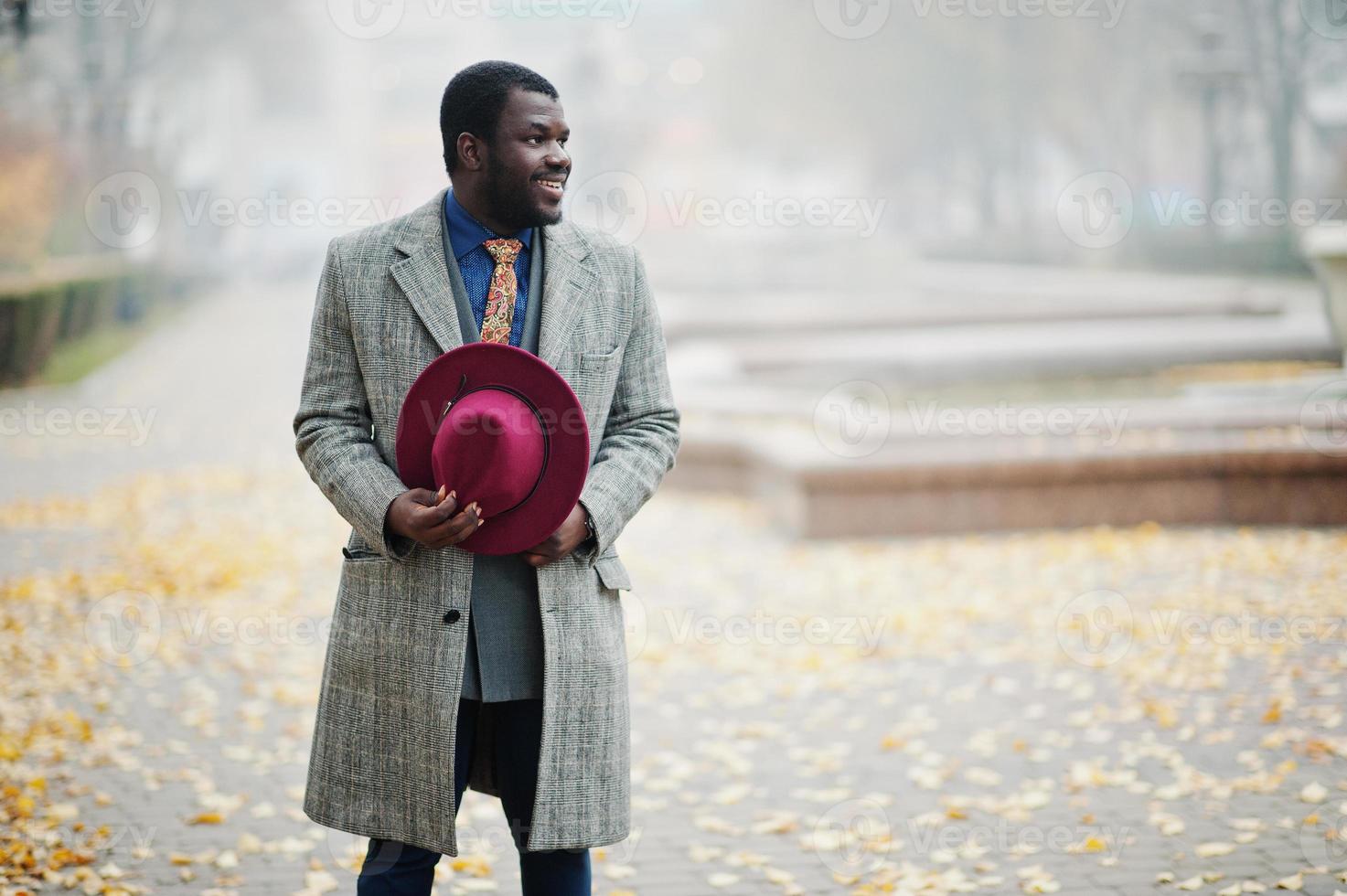 Stylish African American man model in gray coat, jacket tie and red hat posed at foggy weather street. photo
