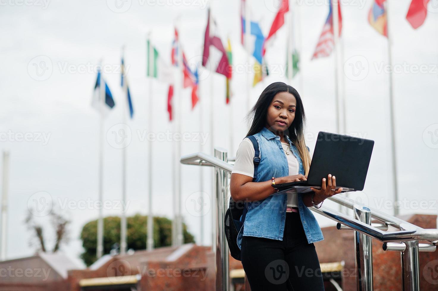 African student female posed with backpack and school items on yard of university, against flags of different countries. She hold laptop on hands. photo