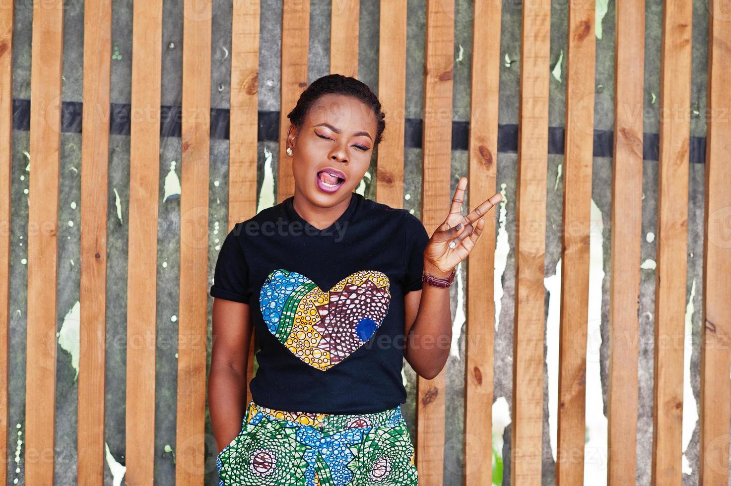 Young african american woman posed against wooden wall, blinking eyes and show two fingers. photo