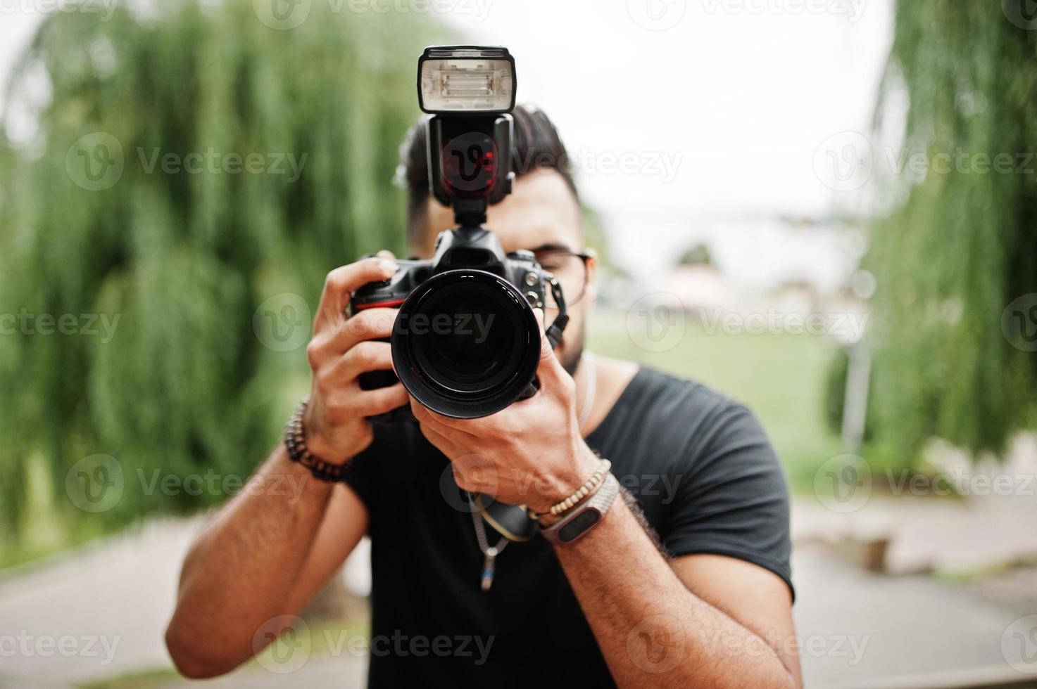 Awesome beautiful tall ararbian beard macho man photographer in glasses and black t-shirt with professional camera at hands. photo