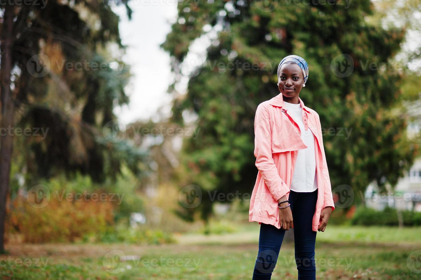 Young modern fashionable, attractive, tall and slim african muslim woman in hijab or turban head scarf and pink coat posed at park. photo