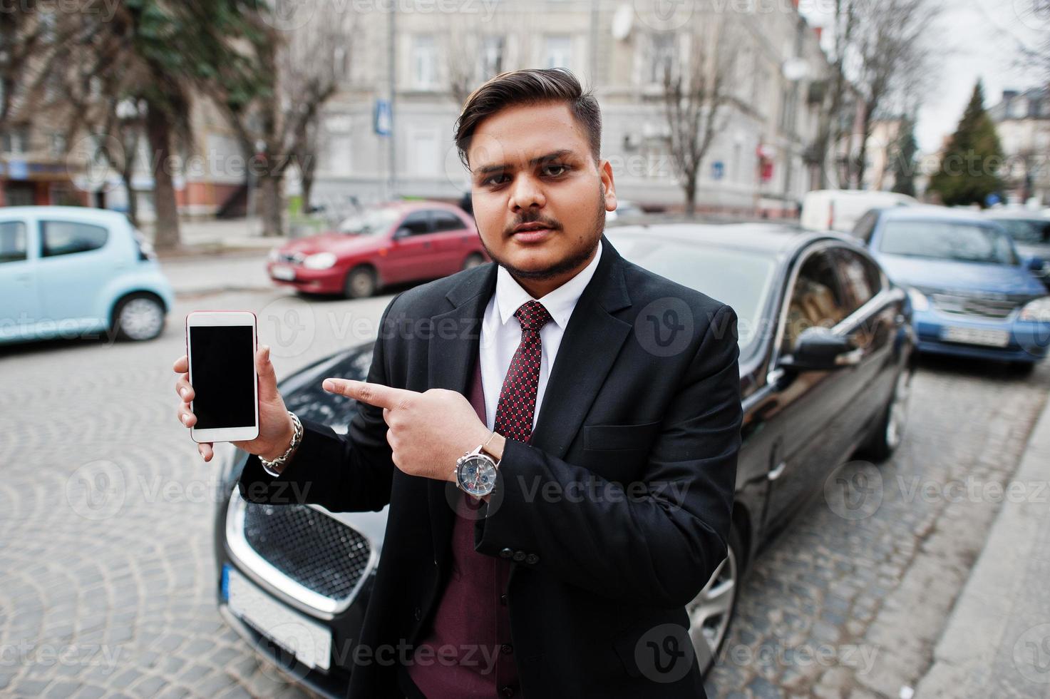 hombre de negocios indio con estilo en ropa formal de pie contra el coche de negocios negro en la calle de la ciudad y muestra el dedo a la pantalla del teléfono móvil. foto