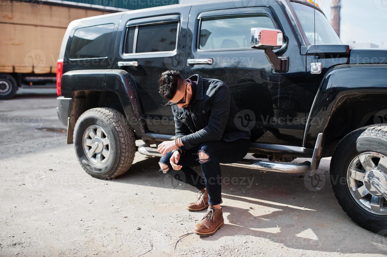 Fashion rich beard Arab man wear on black jeans jacket and sunglasses posed against big black suv car. Stylish, succesful and fashionable arabian model guy. photo