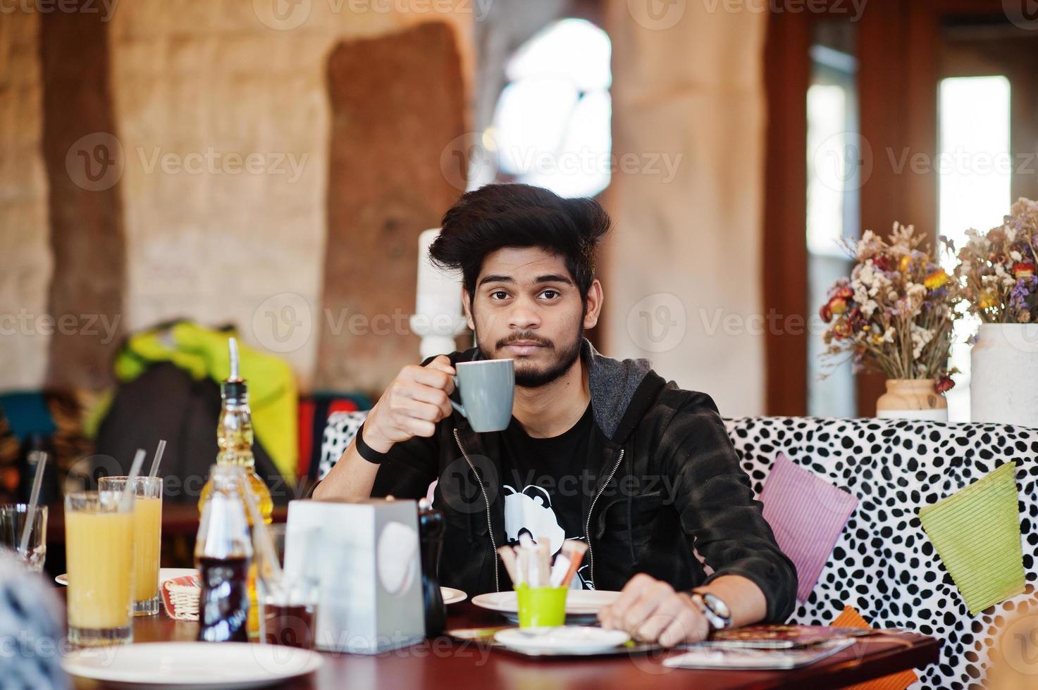 hombre asiático con barba en ropa informal sentado en un café interior y bebiendo café. foto