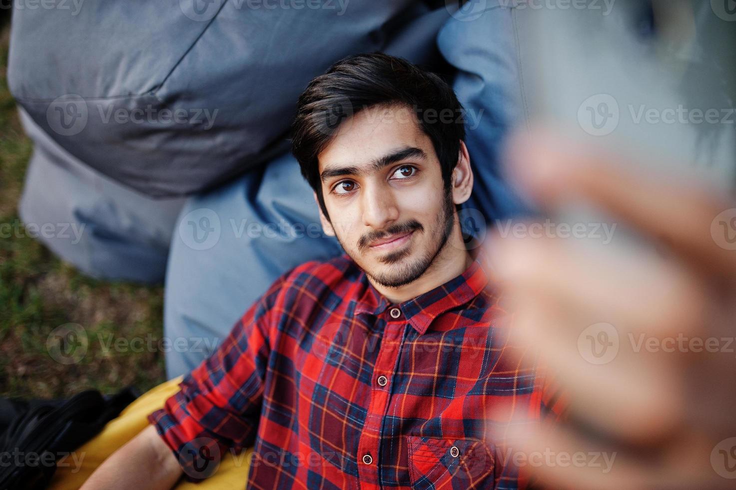 joven estudiante indio con camisa a cuadros y jeans sentado y relajado en almohadas al aire libre y haciendo selfie. foto