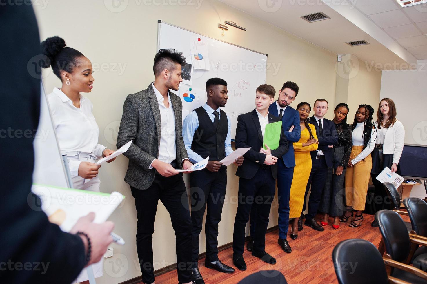 Mixed race business coach presenting report standing near whiteboard pointing on sales statistic shown on diagram and chart teach diverse company members gathered together in conference room. photo