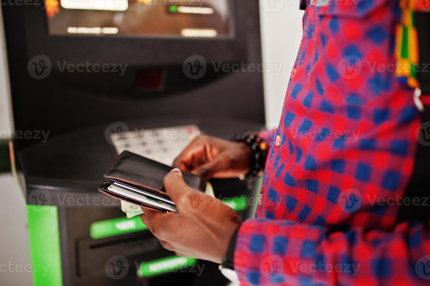 African american man in checkered shirt with backpack holding wallet in hand against atm. Black man traveler. photo