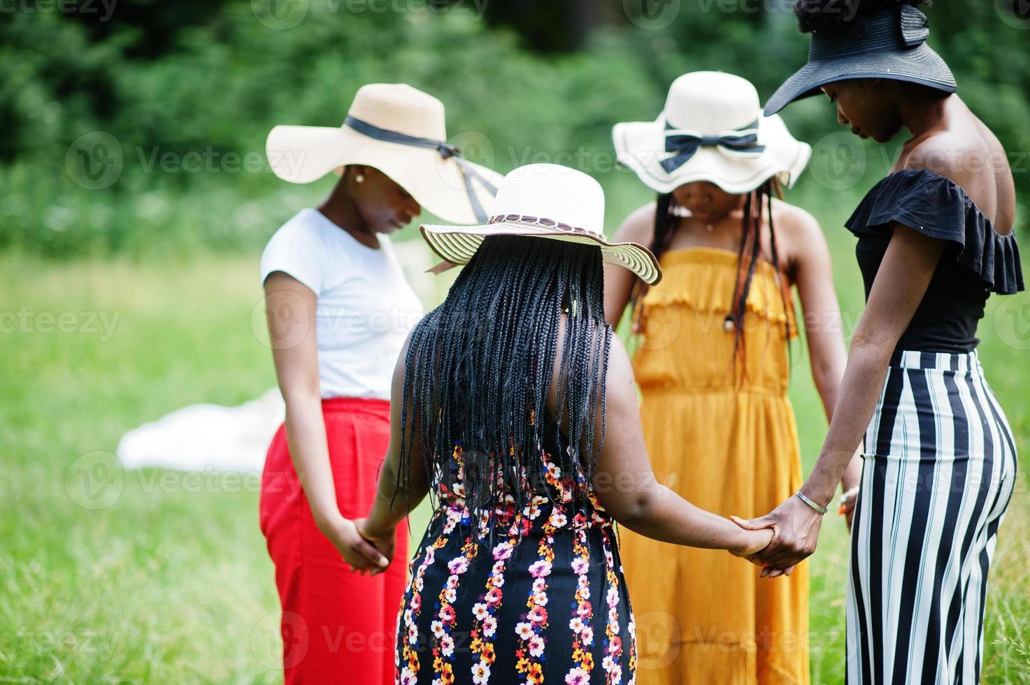 Group of four gorgeous african american womans wear summer hat holding hands and praying at green grass in park. photo