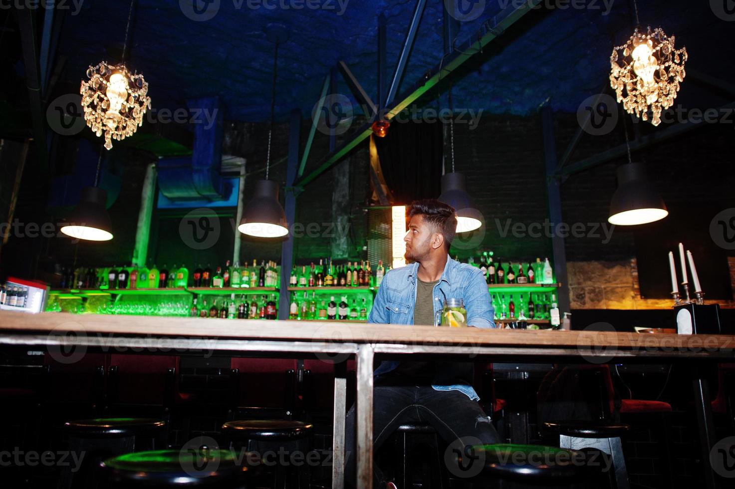 Portrait of handsome successful bearded south asian, young indian freelancer in blue jeans shirt sitting in night club against bar counter with cocktail and having a rest. photo