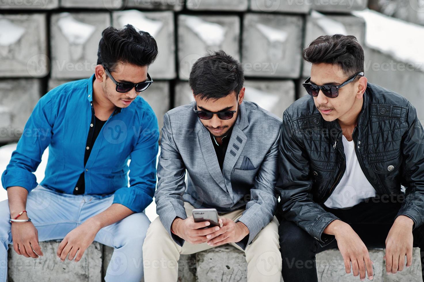 Group of three casual young indian mans in sunglasses posed against stone blocks and looking at mobile phone. photo