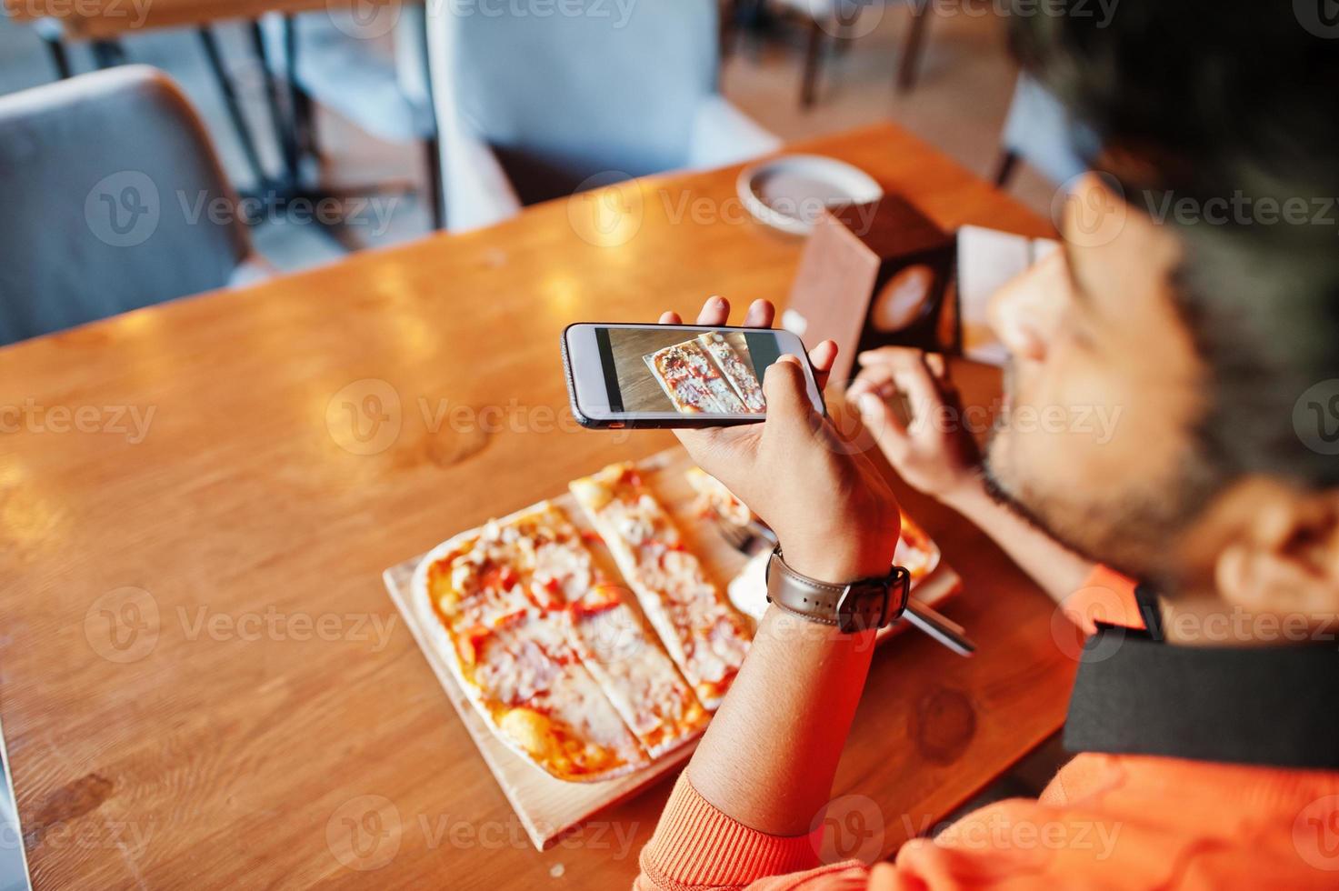 Confident young indian man in orange sweater sitting at pizzeria, eat pizza and makes photo on his phone.