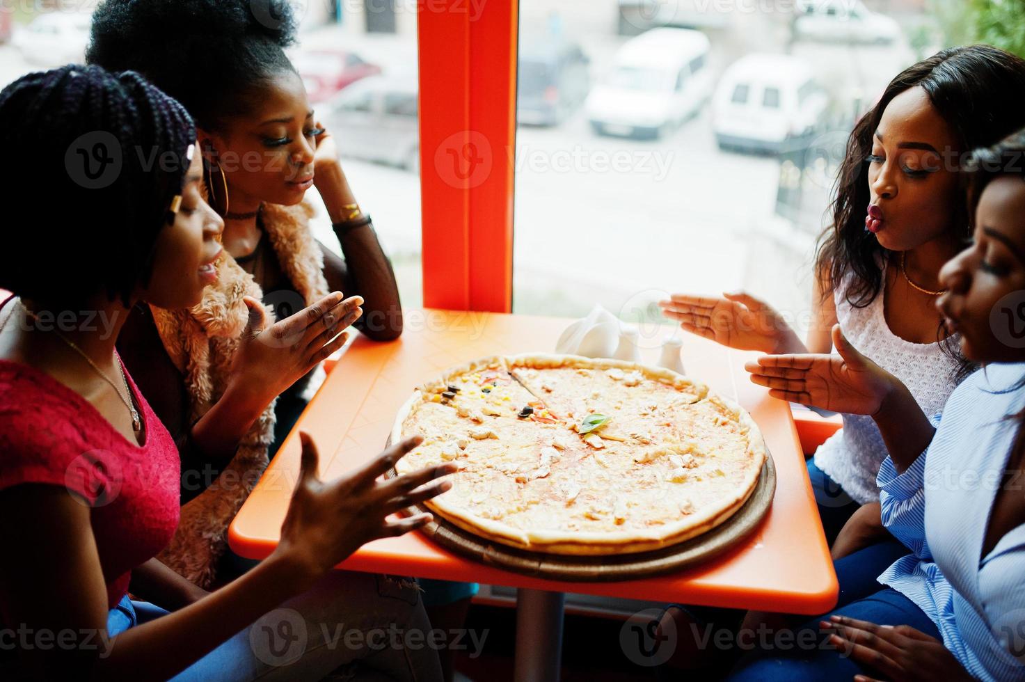 cuatro jóvenes africanas en un restaurante de colores brillantes oliendo pizza y divirtiéndose juntas. foto