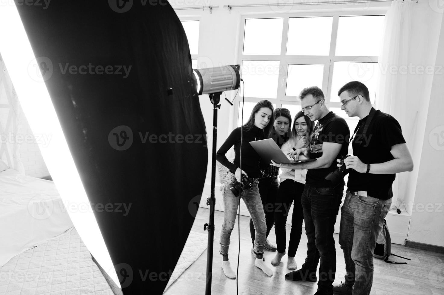 Photographer explaining about the shot to his team in the studio and looking on laptop. Talking to his assistants holding a camera during a photo shoot. Teamwork and brainstorm.