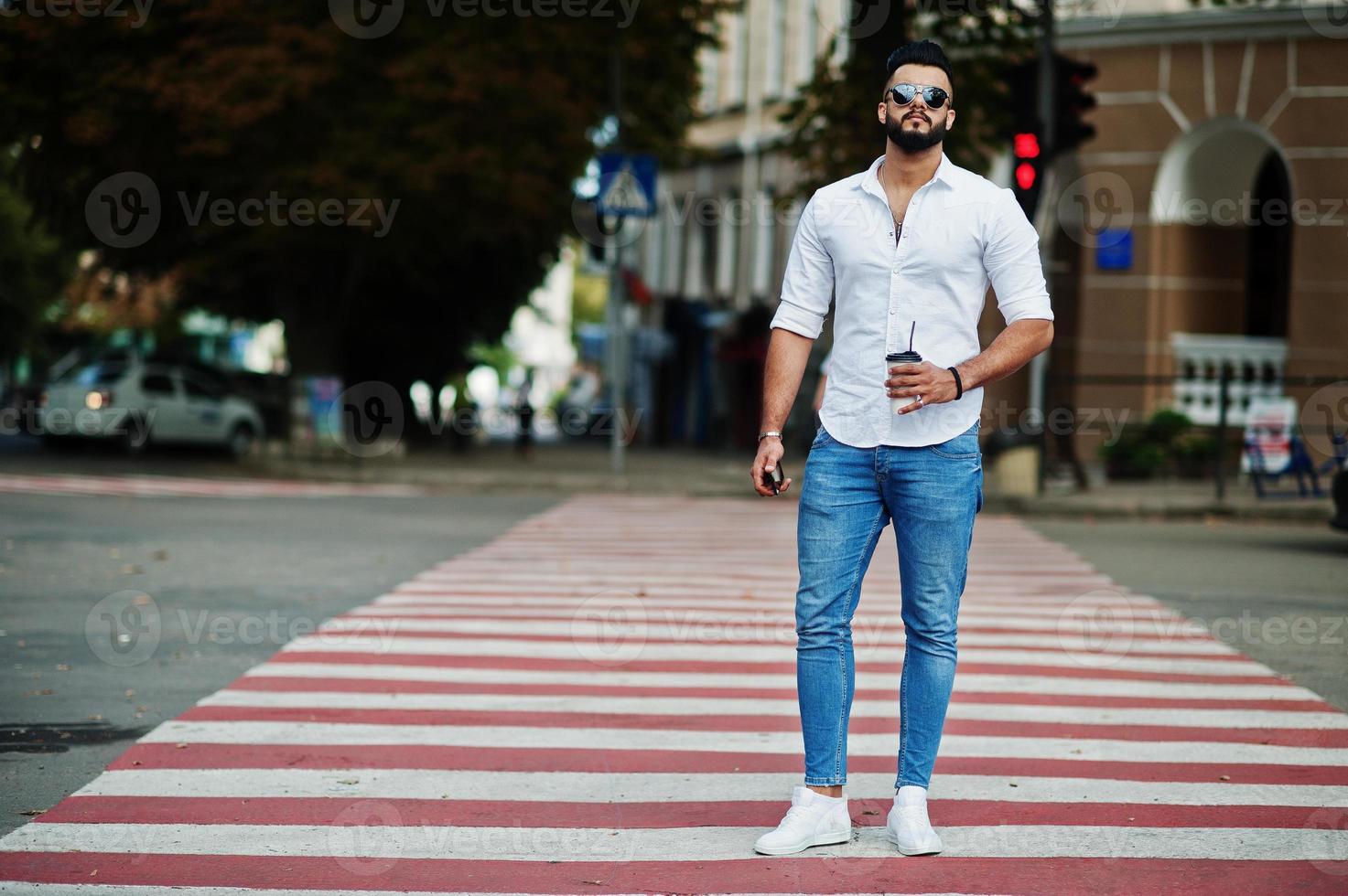 Elegante modelo de hombre árabe alto con camisa blanca, jeans y gafas de sol en la calle de la ciudad. chico árabe atractivo con barba con una taza de café caminando por un paso de peatones o un paso de peatones. foto