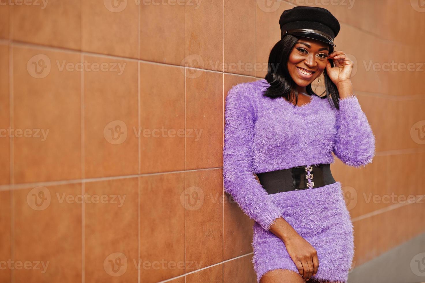 mujer afroamericana con vestido violeta y gorra posada al aire libre contra la pared naranja. foto