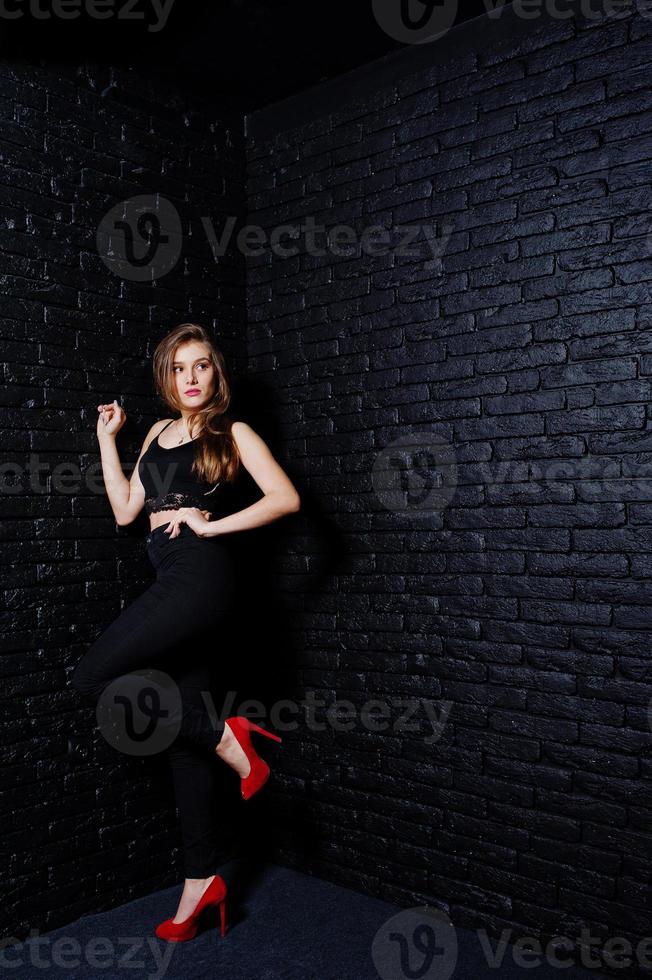 Handsome brunette girl wear on black and red high heels, posing at studio against dark brick wall. Studio model portrait. photo