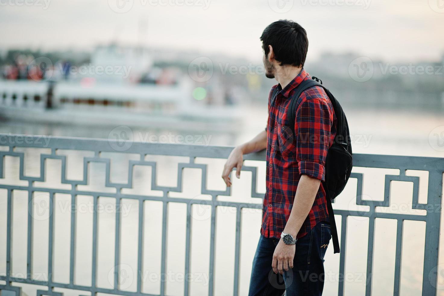 Young indian student man at checkered shirt and jeans with backpack posed on evening city against lake and boat. photo