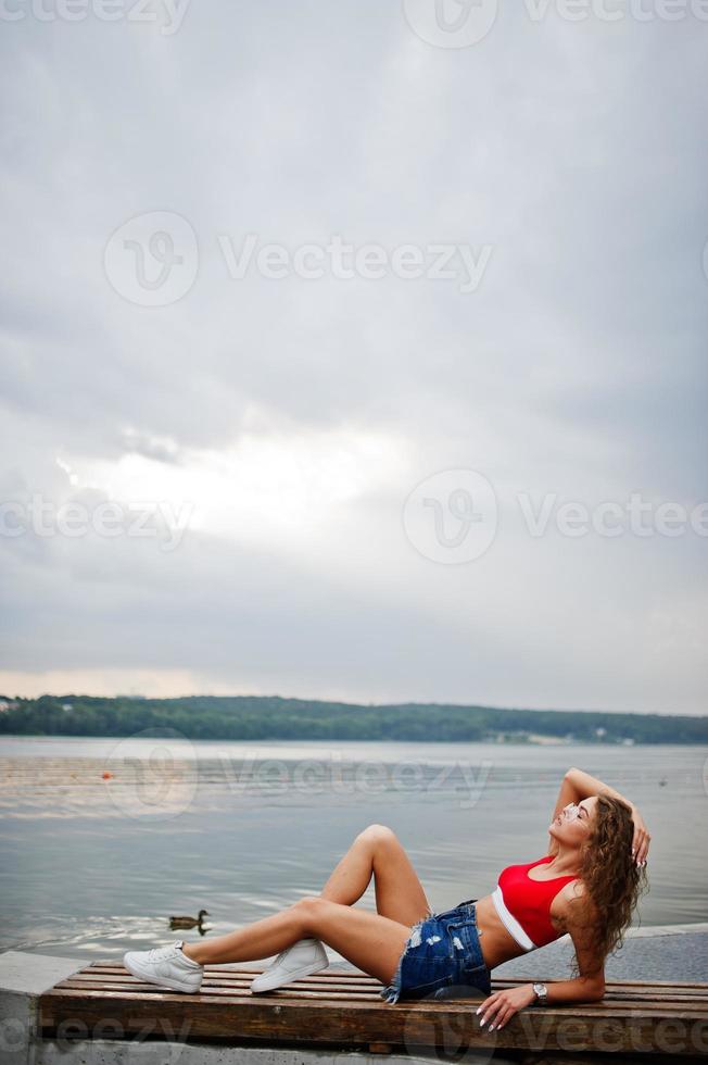 chica sexy modelo rizada en top rojo, pantalones cortos de mezclilla, anteojos y zapatillas posadas en un banco contra el lago. foto