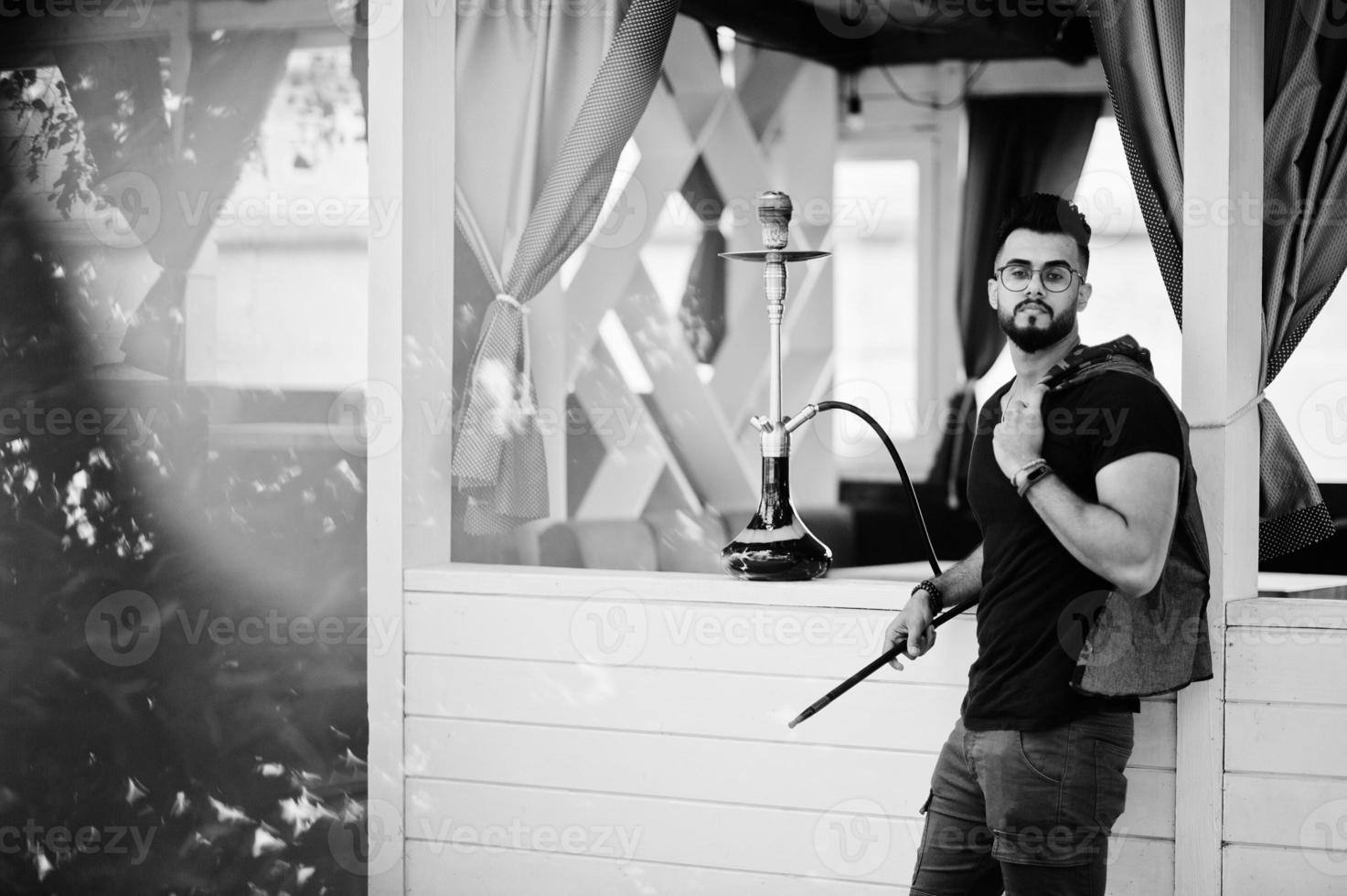 hombre árabe de barba elegante con gafas y camiseta negra fumando narguile al aire libre. modelo árabe descansando. foto