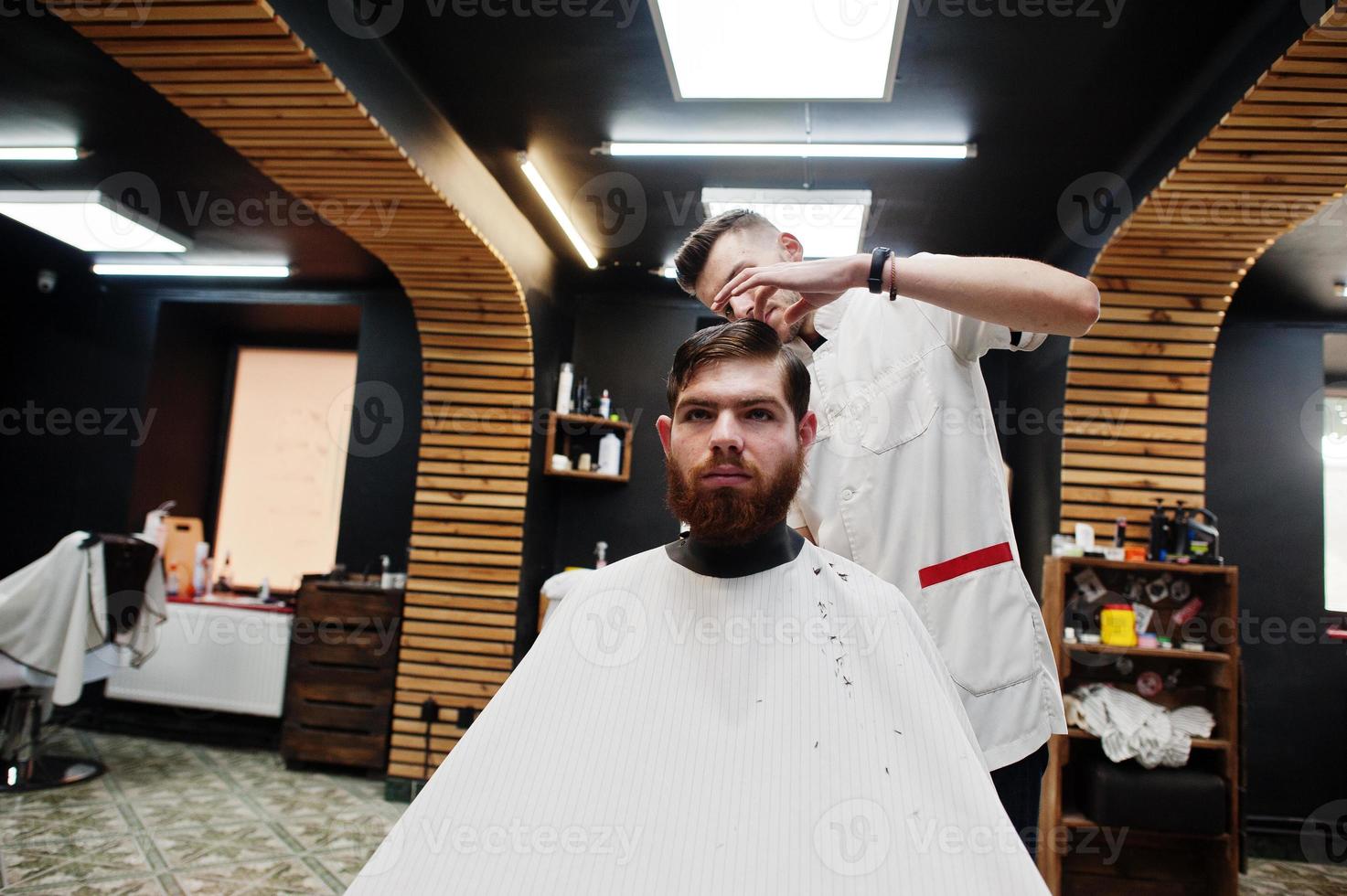 Young bearded man getting haircut by hairdresser while sitting in chair at barbershop. Barber soul. photo